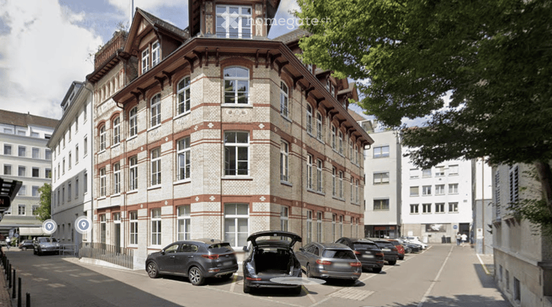 A multi-story brick building with large windows and decorative architectural details, situated on a street with parked cars and trees.
