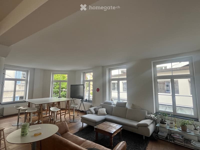 Bright living room with large windows, grey sectional sofa, coffee table, small round table, stools, and a TV screen on a stand.