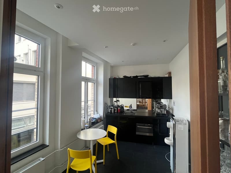 Modern kitchen with black cabinets, stainless steel appliances, and a small dining area with yellow chairs and a white table. The space features large windows and a sleek, contemporary design.
