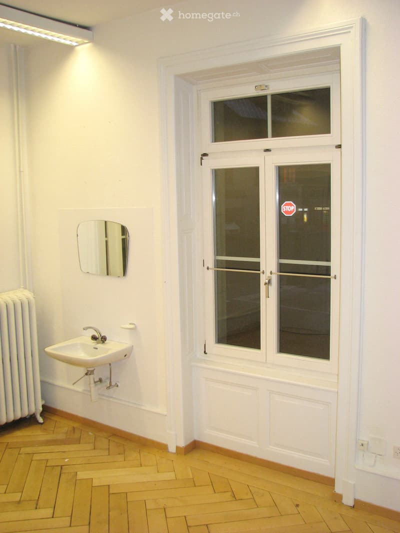 Small bathroom with wooden floor, white walls, a sink, a mirror, and a radiator. A window with frosted glass panes is visible.