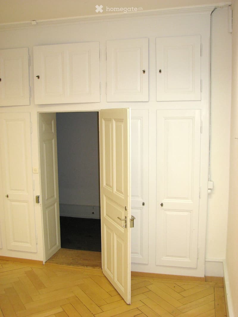 Empty hallway with wooden flooring, white walls, and multiple white doors, one of which is open to reveal a dark room beyond.
