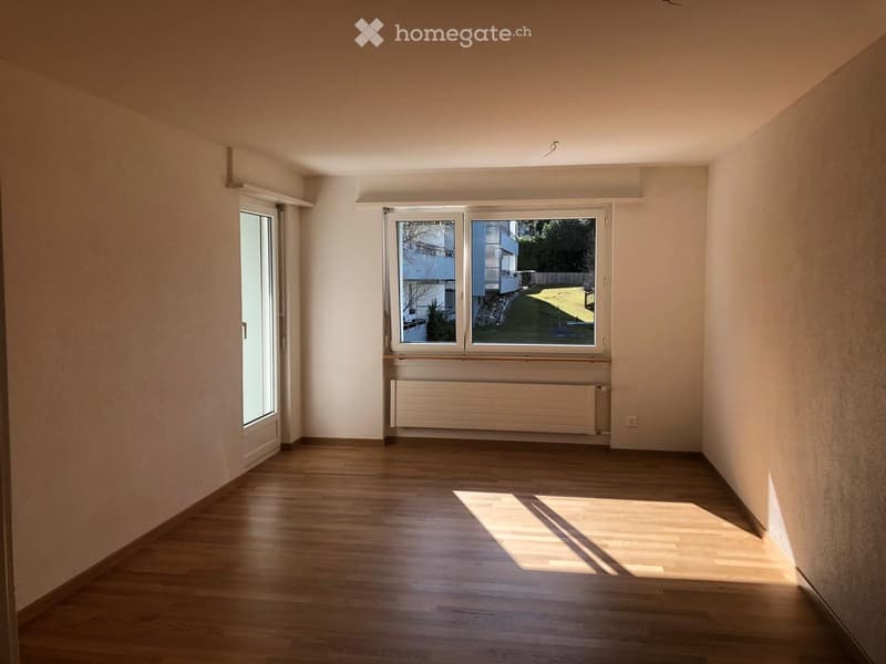 Empty room with white walls, hardwood floors, large window with view of lawn and trees, white radiator