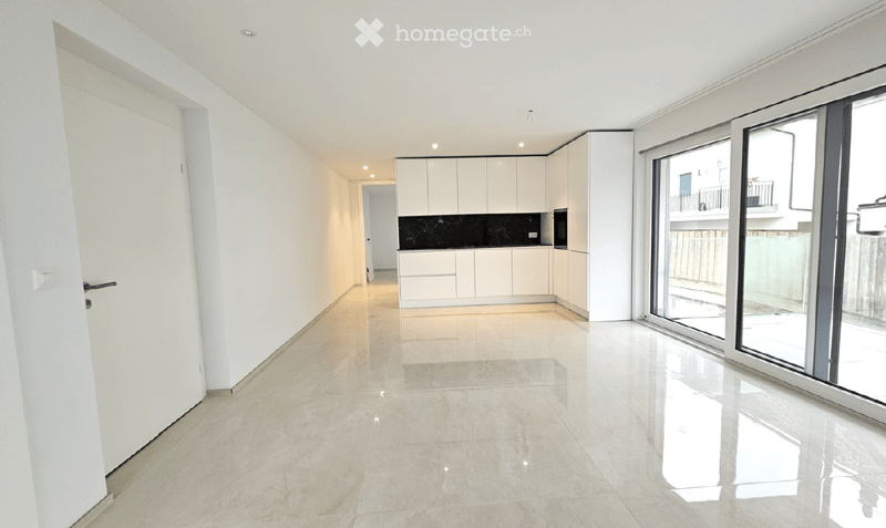 Bright, open-plan space with white walls, polished tile flooring, and a modern white kitchen area. Large glass sliding doors lead to a balcony.