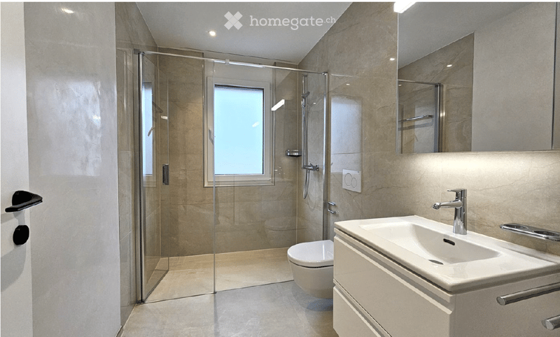 Modern bathroom with a glass-enclosed shower, a white sink with a chrome faucet, a toilet, and a large mirror above the sink. The walls are tiled in a neutral tone, and the floor is light-colored.