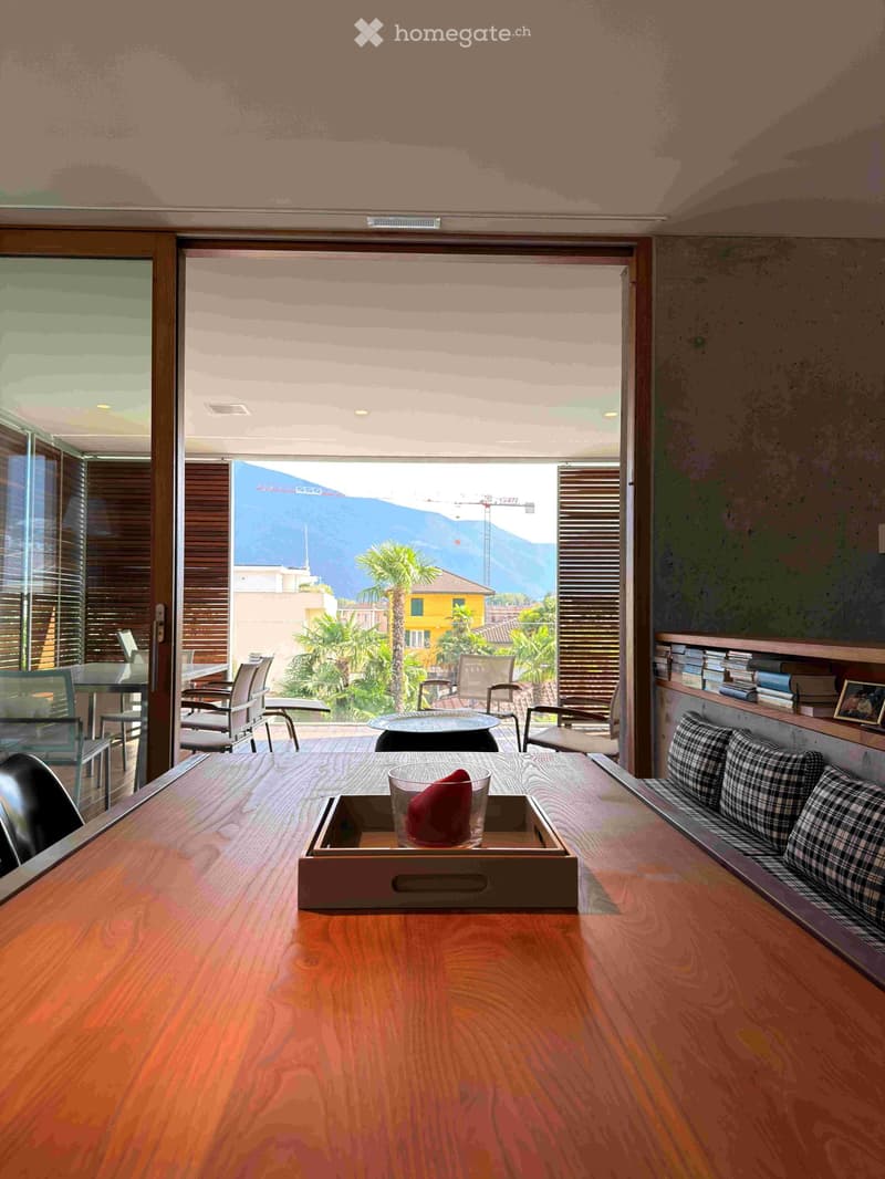 Modern living room with large windows offering a scenic mountain view, featuring a wooden table, chairs, a sofa, and a bookshelf with books and a picture frame.