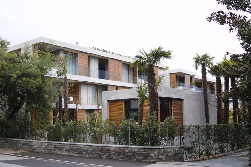 Modern two-story building with concrete and wood accents, large windows, balconies, and surrounded by tall palm trees and greenery.