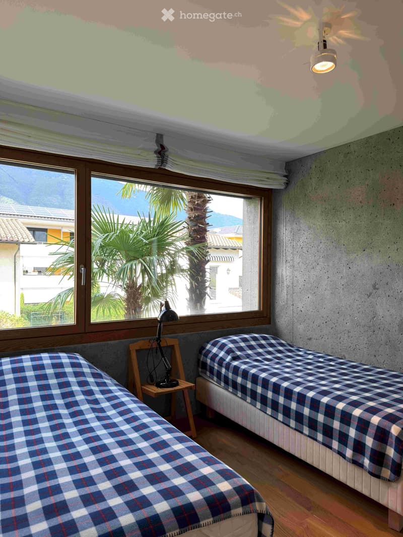 A modern bedroom with two single beds, each covered in blue and red checkered blankets, a wooden nightstand with a black lamp, and large windows offering a view of palm trees and mountains.
