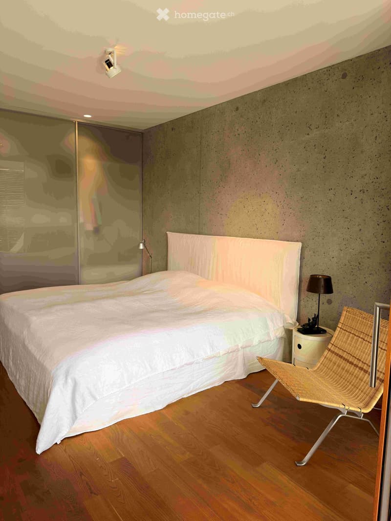 Modern bedroom with a large bed, white bedding, a wicker chair, a nightstand with a lamp, and a sleek headboard. The room features exposed concrete walls and wooden flooring.