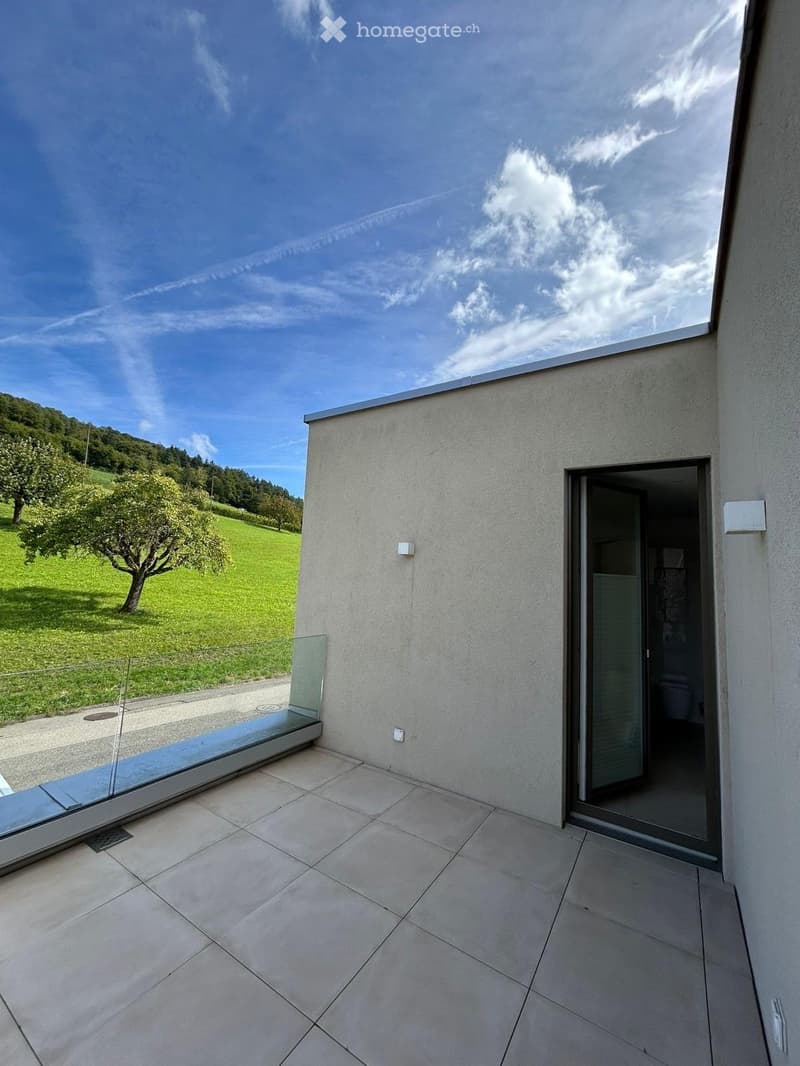 Modern balcony with tiled floor, glass railing, and open door leading to interior space. Scenic view of green landscape and tree.