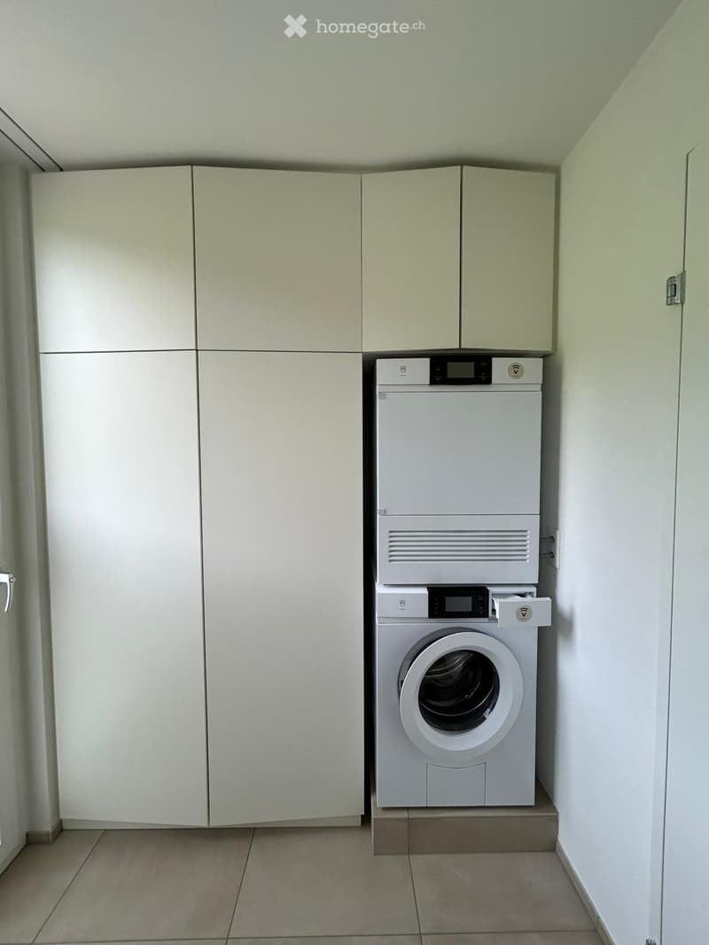 A small, well-lit room with a washing machine and dryer stacked vertically, surrounded by light-colored cabinets.