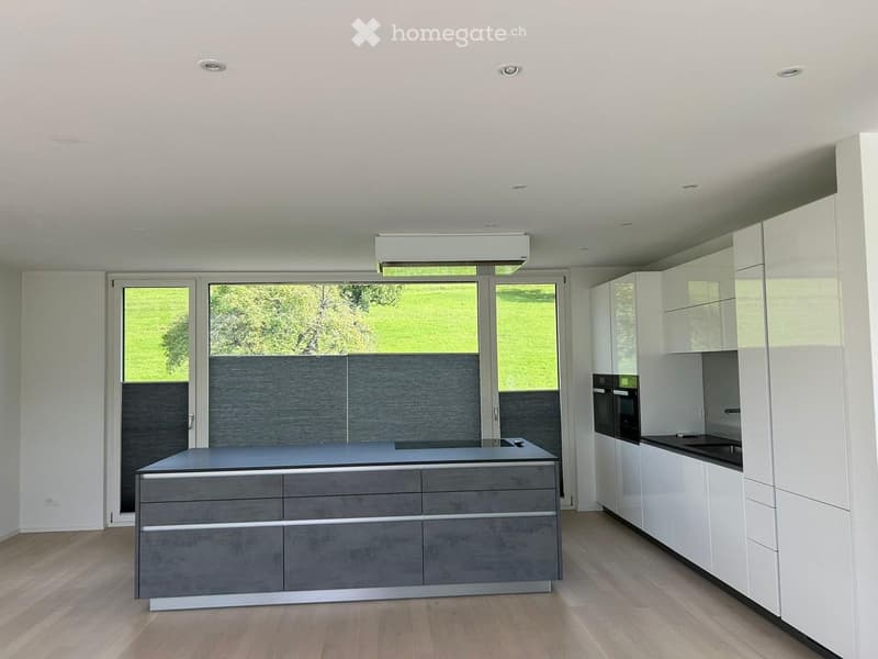 Modern kitchen with white cabinets, grey island, and large windows overlooking a green area.