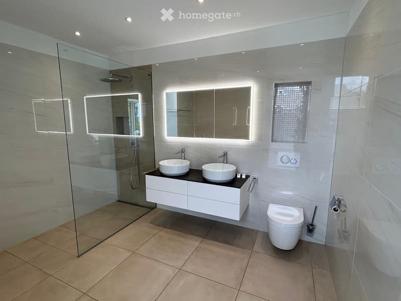 Modern bathroom with white tiles, a double sink vanity, a toilet, a large mirror with LED lighting, and a glass-enclosed shower area.