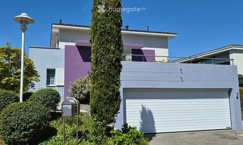 Modern two-story house with white and purple exterior, large garage, and well-maintained landscaping including a tall evergreen tree and shrubs.