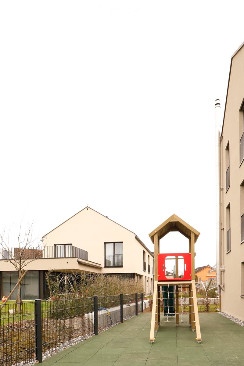 modern apartment complex with a playset and green flooring, a playground, black fence, buildings on the side, a tree, small plants, and a small garden in the corner