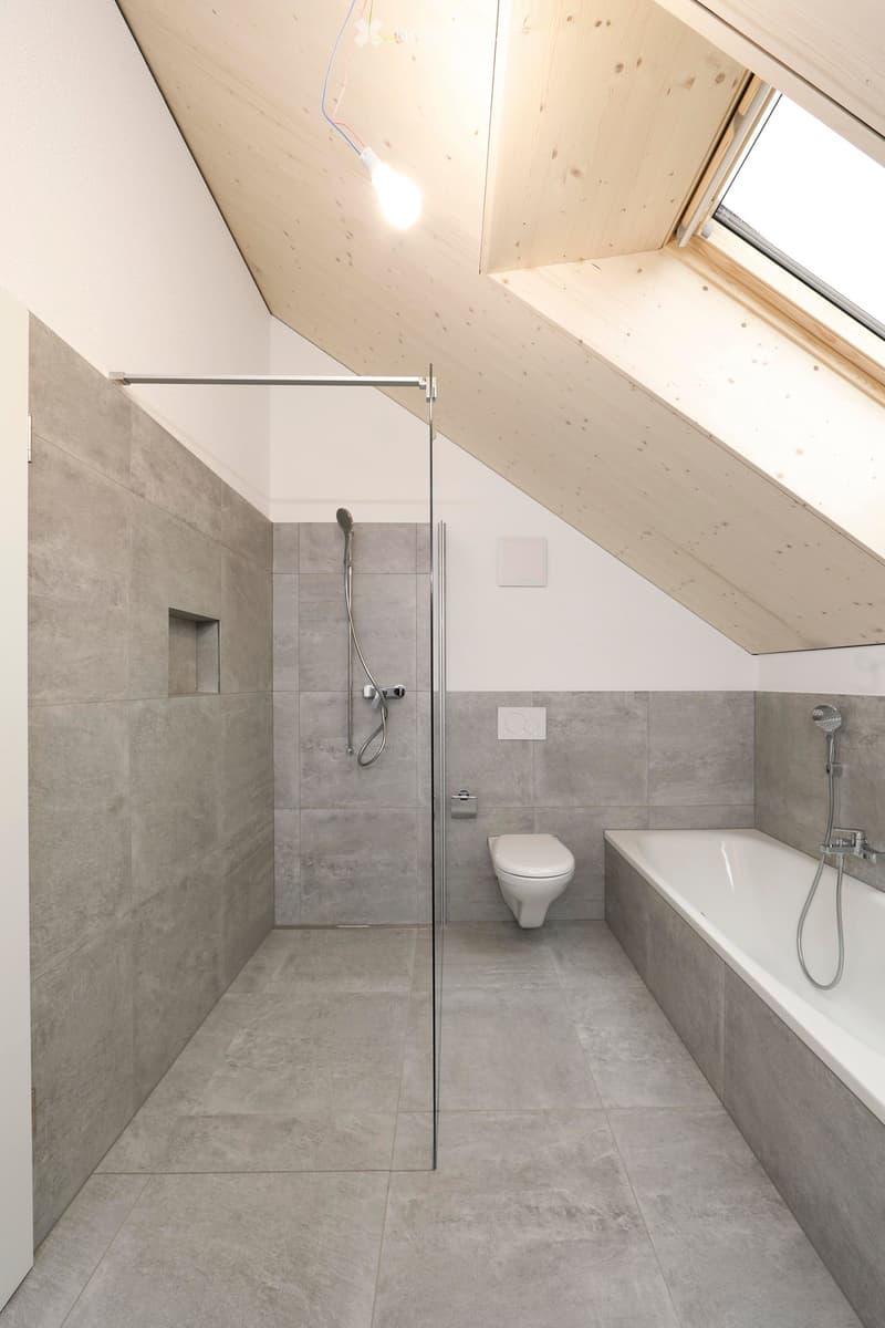 A minimalist attic bathroom with gray tiles, a shower, bathtub, toilet, and exposed wooden ceiling.