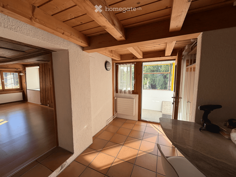 Empty room with wooden ceiling beams, tiled floor, and large windows. No furniture or equipment present.