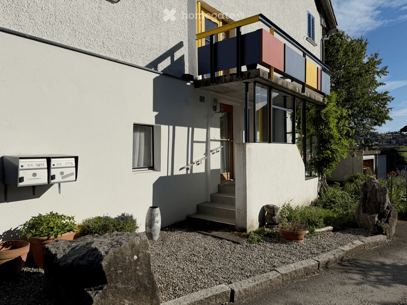 Two-story building with white facade, balcony, and small garden area with potted plants and decorative rocks.