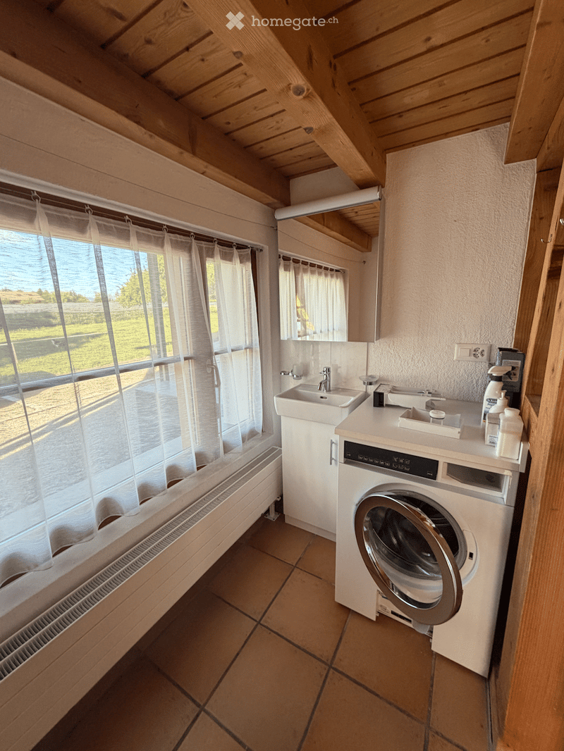Bathroom with wooden ceiling, tiled floor, sink, washing machine, and large window with curtains.