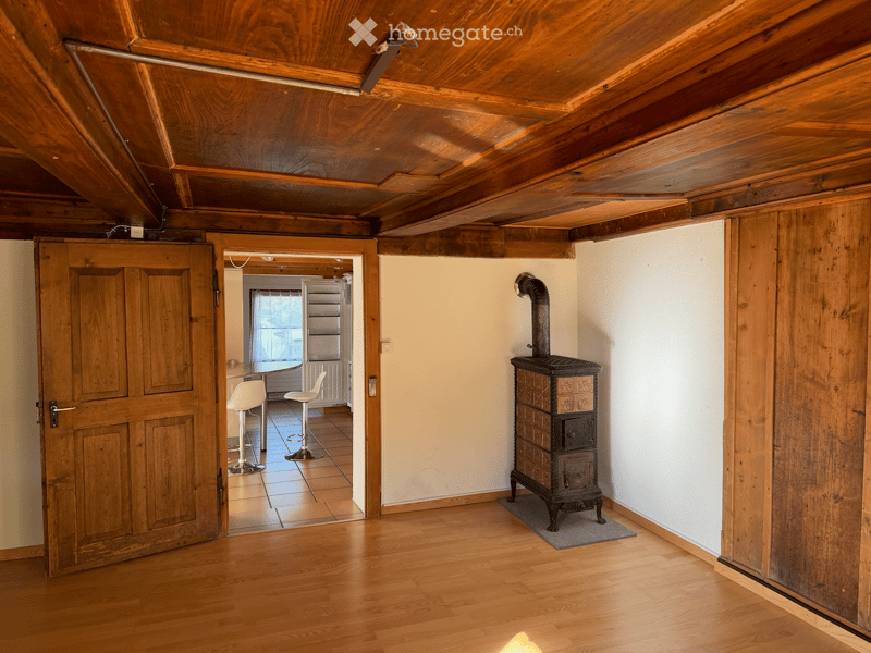 The room features exposed wooden beams on the ceiling, wooden flooring, and a wooden stove. A doorway leads to another room with two white stools and a small table.