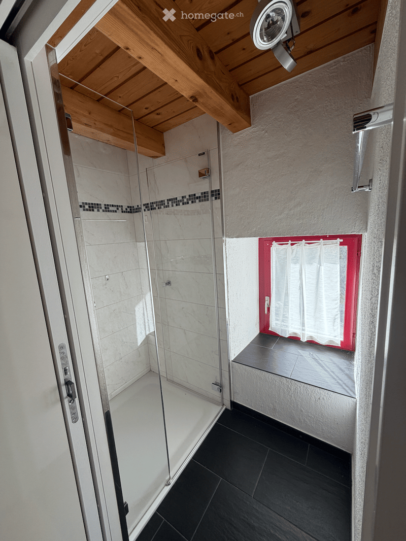 Modern bathroom with a glass-enclosed shower, black tiled floor, white walls, and a small window with a red frame and white curtain.
