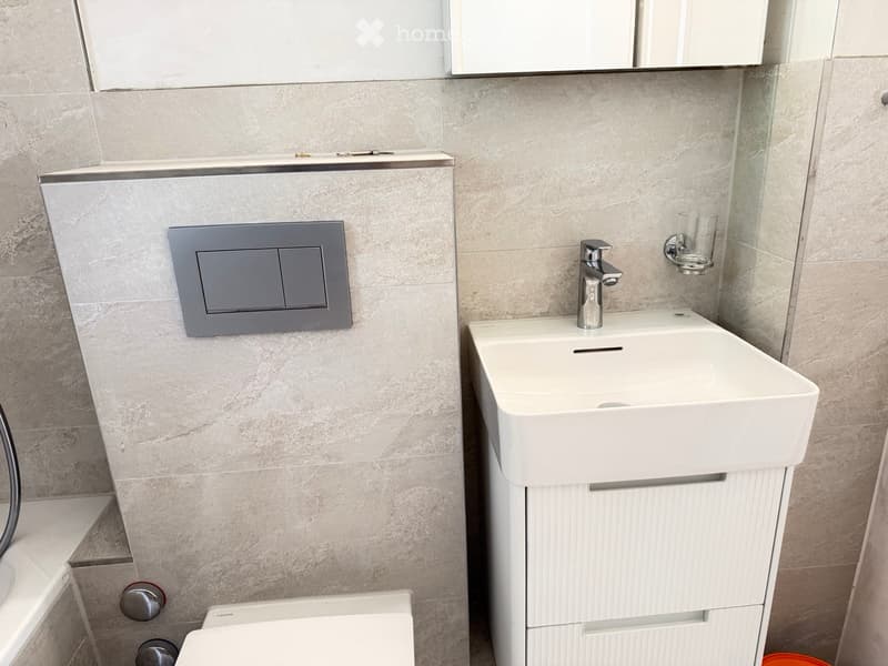 Modern bathroom with a white sink, chrome faucet, and a mirror above. The walls are tiled in a light, marble-like pattern.