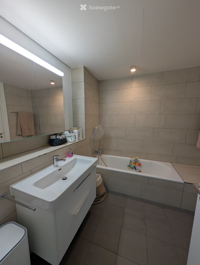 Modern bathroom with a white sink, bathtub, and large mirror. The room features beige tiles and a sleek design.