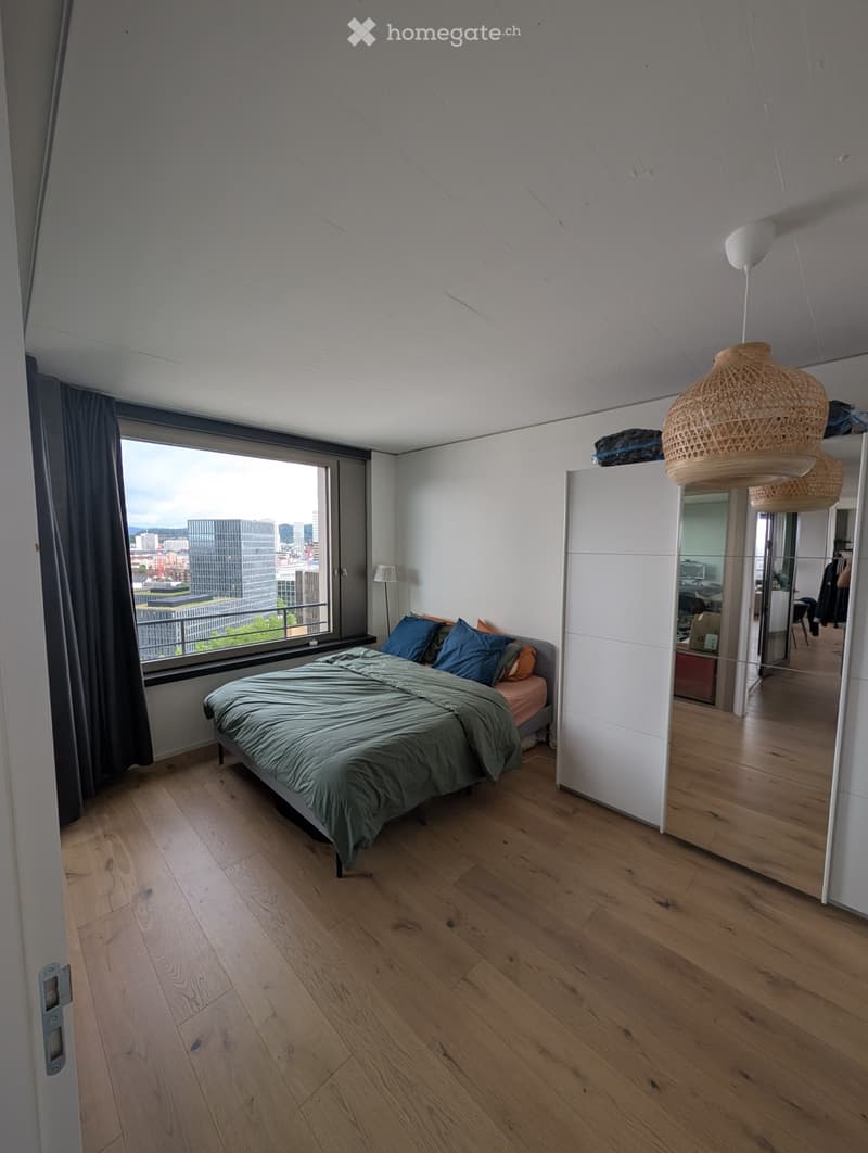 Modern bedroom with wooden flooring, large window offering city view, bed with green duvet and blue pillows, hanging woven lamps, and a mirrored wardrobe.