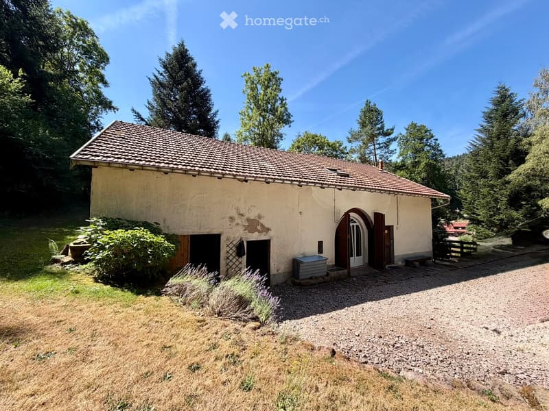 A small, rustic house with a tiled roof and beige walls, surrounded by greenery and a gravel driveway.