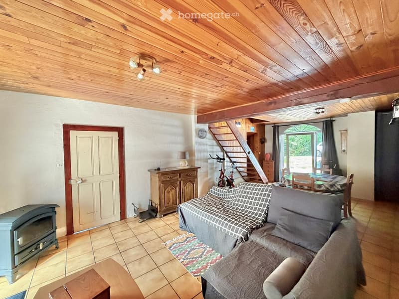 Living room with exposed wooden ceiling beams, tiled floor, sofa, coffee table, fireplace, dining area with table and chairs, and a staircase leading to an upper level.