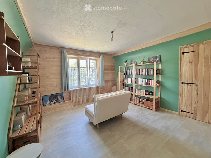 Cozy living room with light green walls, wooden shelving, a cream-colored sofa, and a large window with curtains. The room features a bookshelf filled with books and decorative items, a picture frame on the wall, and a radiator beneath the window.