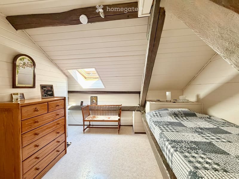 Cozy attic bedroom with slanted wooden ceilings, a single bed with a patterned quilt, a wooden dresser, a small wooden chair, and a skylight window allowing natural light.