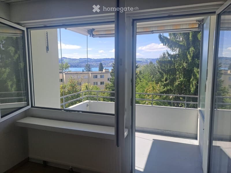 Balcony with large windows offering scenic views of trees, water, and distant hills.