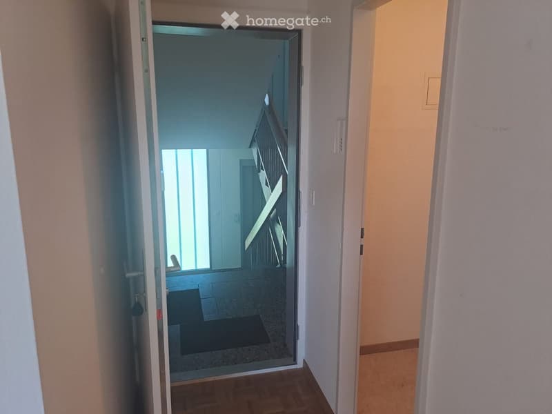 A narrow hallway with light-colored walls and wooden flooring, leading to an open doorway that reveals a glimpse of a modern interior space with a staircase and glass walls.