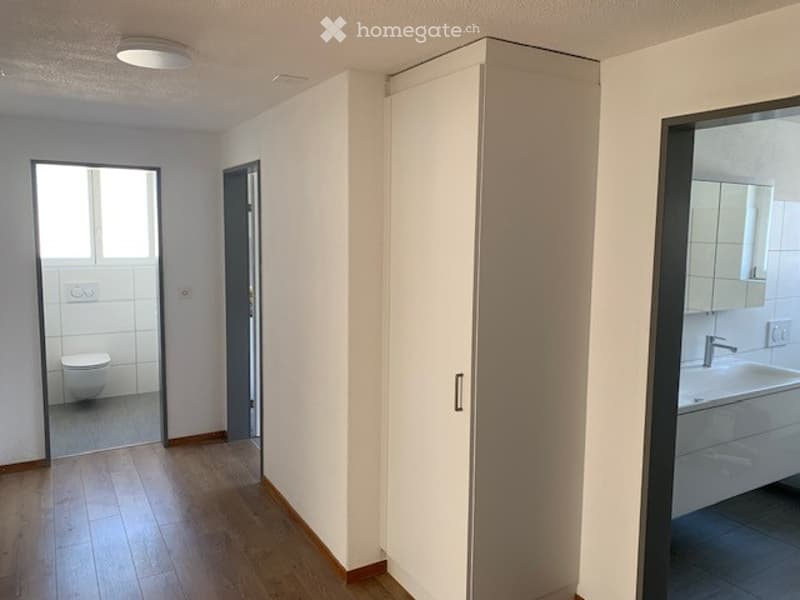 Empty hallway with wooden flooring, white walls, and a tall white cabinet. A partially visible bathroom with a toilet and sink is seen through an open doorway.