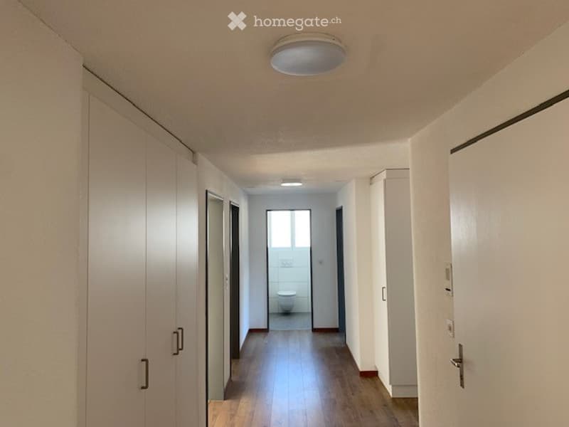 Empty hallway with wooden flooring, white walls, and a single ceiling light fixture. A small bathroom is visible at the end of the corridor.