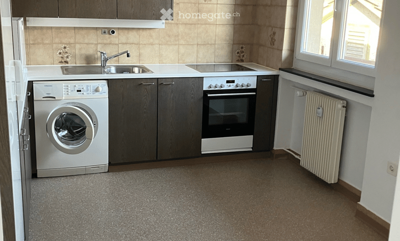 Modern kitchen with a washing machine, oven, sink, faucet, and radiator. The space features wooden cabinets, tiled walls, and a grey carpeted floor.