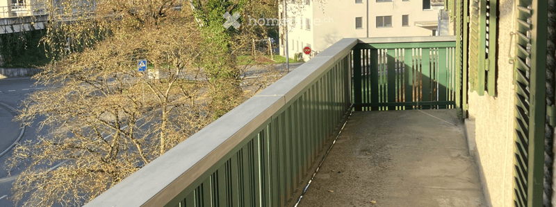 Balcony with green railing, overlooking a street with trees and a building in the background.