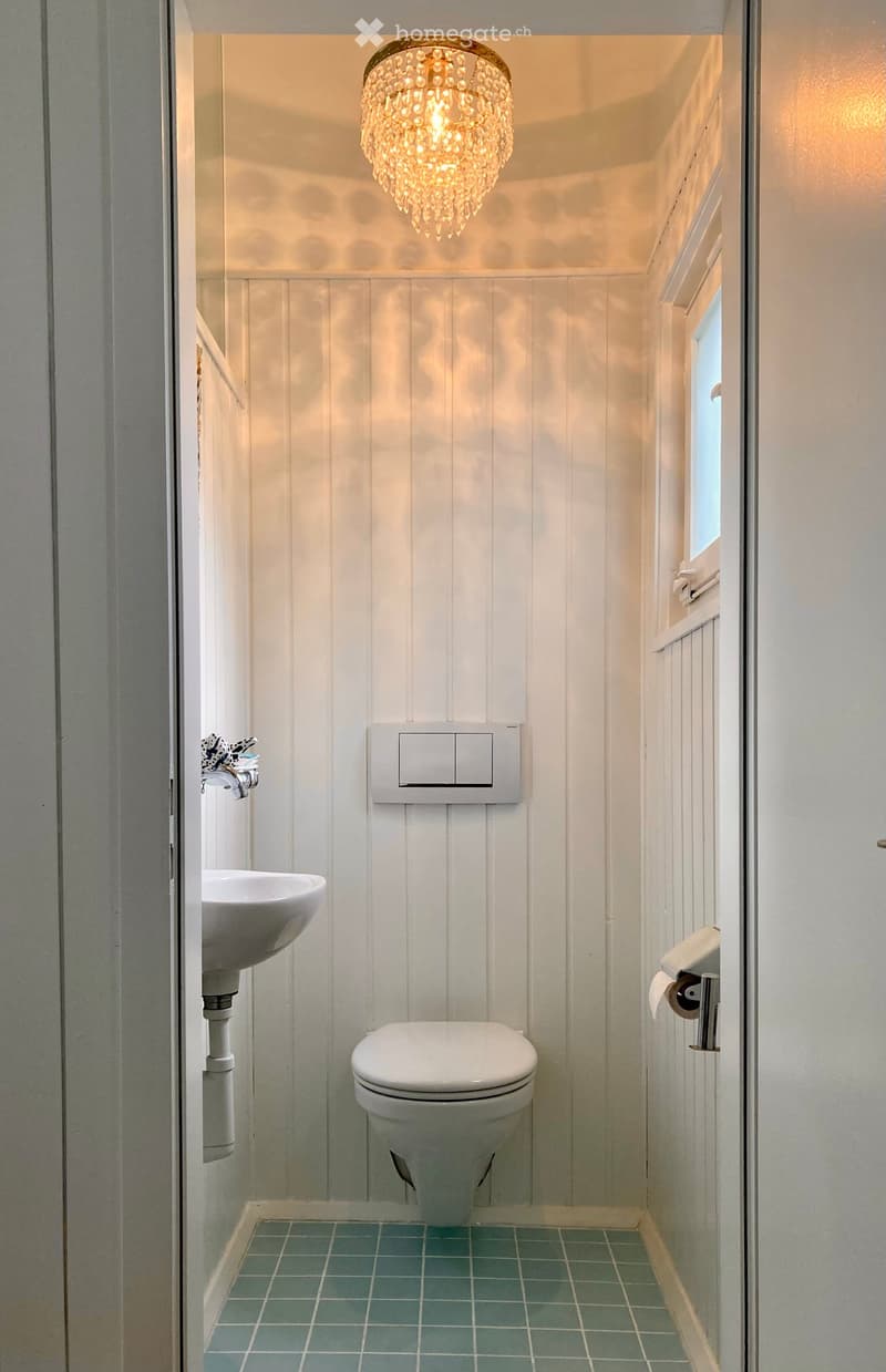Compact bathroom with white paneled walls, teal tiled floor, a toilet, a sink with a modern faucet, and a decorative chandelier light fixture.