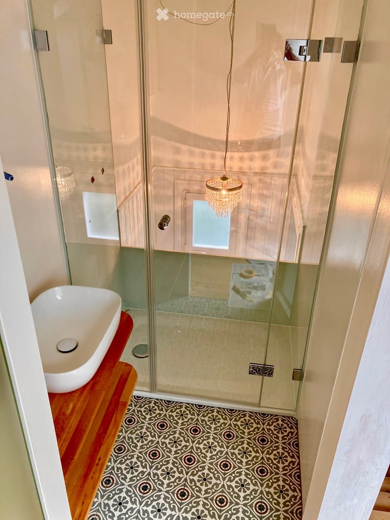 Elegant bathroom with a white bathtub on a wooden platform, a glass-enclosed shower with a chandelier, and patterned tiles on the floor.