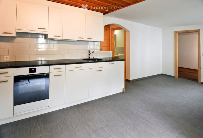 Modern kitchen with white cabinets, black countertops, and stainless steel appliances. Features a sink with a faucet and an oven, with a grey tiled floor and a wooden ceiling.