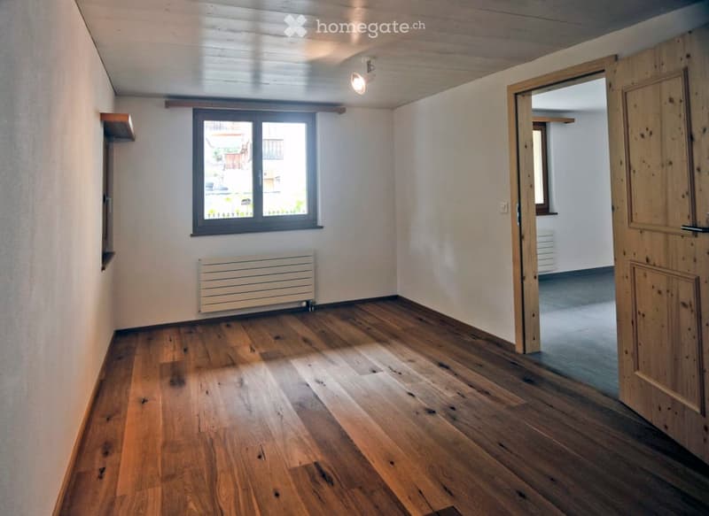 Empty room with wooden flooring, white walls, a window with a view, a radiator, and a wooden door leading to another room.