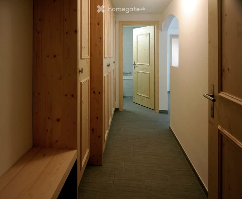 A narrow hallway with wooden panel walls, a tiled floor, and several wooden doors leading to rooms. The hallway features a bench on the left side and an arched opening on the right.