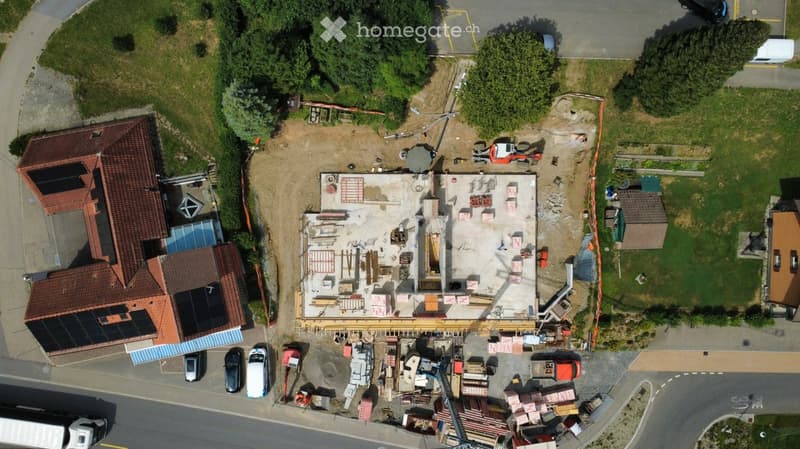 Aerial view of a residential area showing a partially constructed building with exposed concrete and construction materials, surrounded by other houses and greenery.