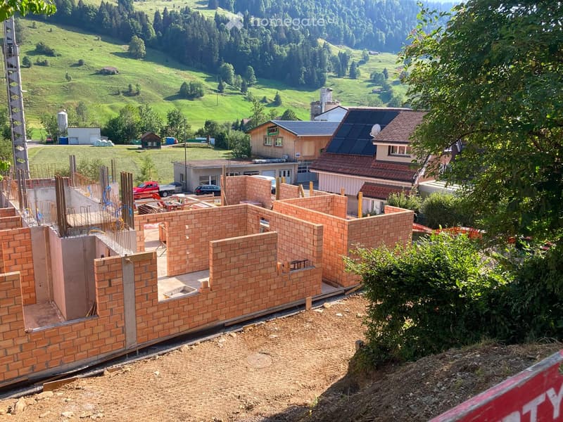 Under construction house with exposed brick walls and a raised ground floor, surrounded by greenery and other buildings in a rural landscape.