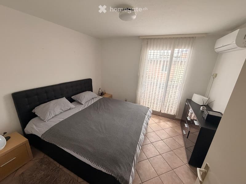 A bed with grey pillows and a blanket, nightstands on either side, wooden cabinet with drawers, air conditioner, white curtains on the balcony