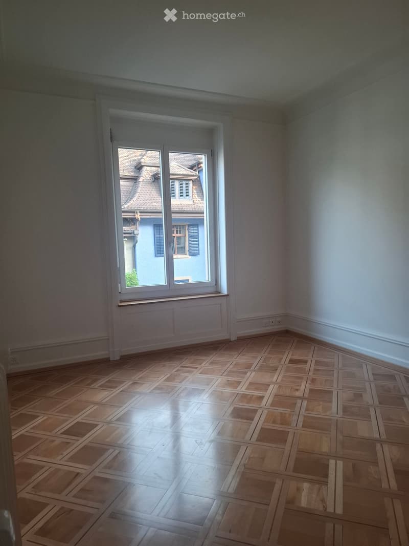 Empty room with wooden parquet flooring and large window showing a view of neighboring houses.