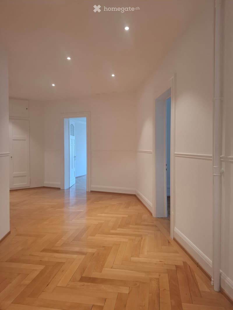 Empty hallway with light wooden herringbone flooring, white walls, and recessed ceiling lights. Two open doorways lead to other rooms.