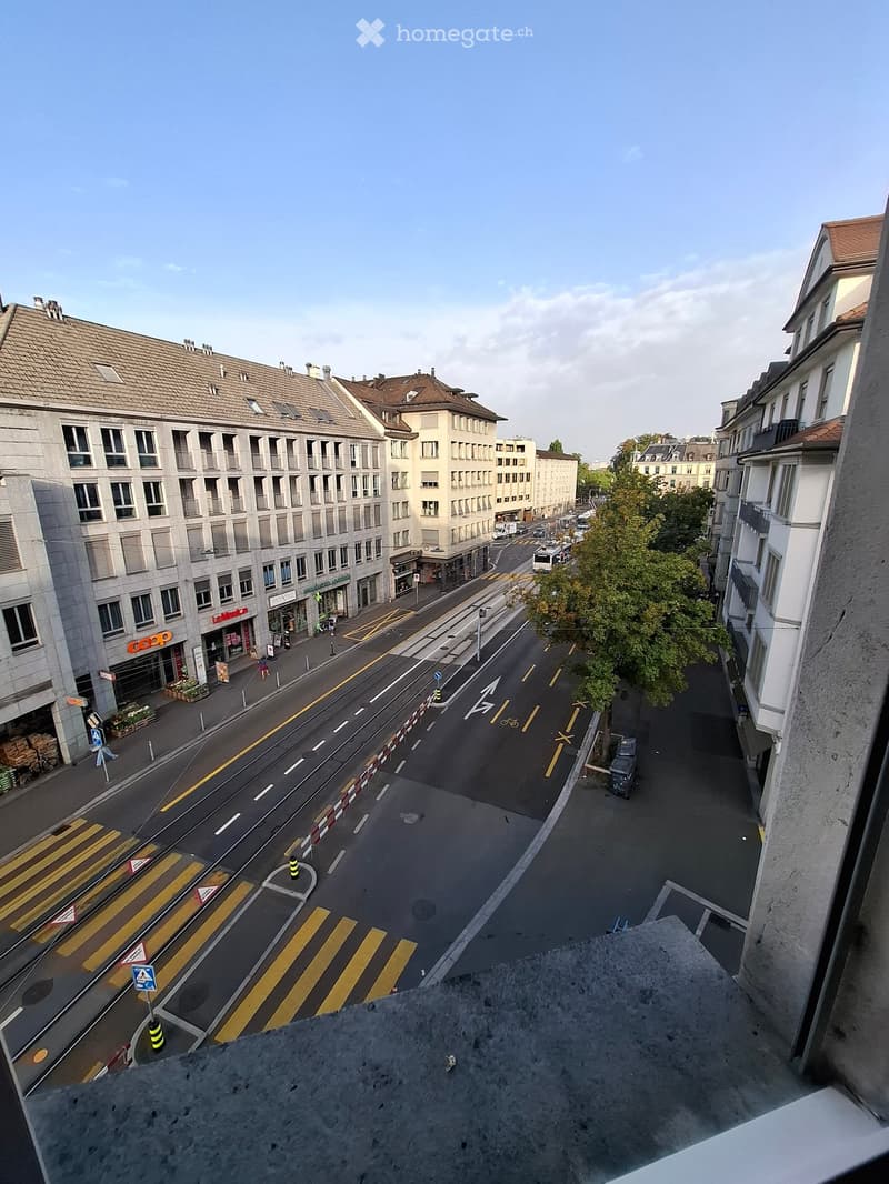 View from a balcony overlooking a quiet urban street with crosswalks, traffic markings, and buildings on both sides.