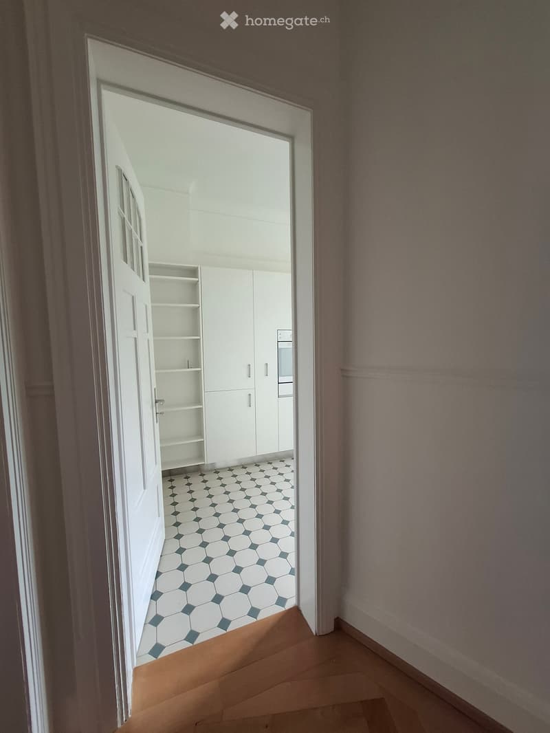 A narrow hallway with wooden flooring leading to a room with white cabinets and a hexagonal tile floor.