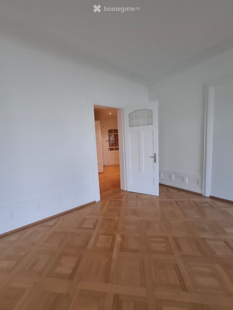 Empty hallway with wooden flooring and white walls, featuring a closed white door with glass panels and an open doorway leading to another room.
